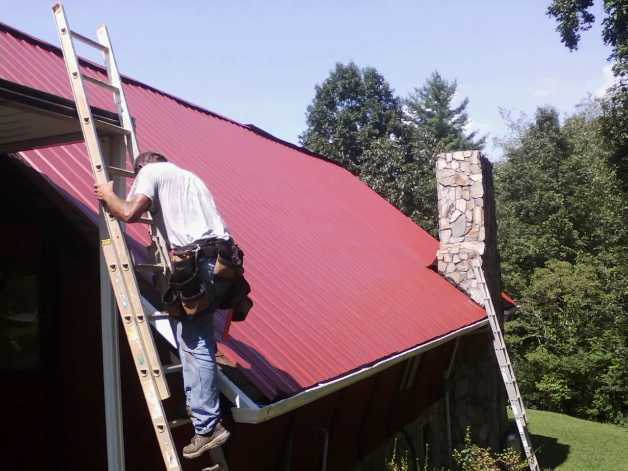 Roofer on a ladder beside a red metal roof and stone chimney — Soffit & Fascia Repair by Roofing Brothers in Bergen